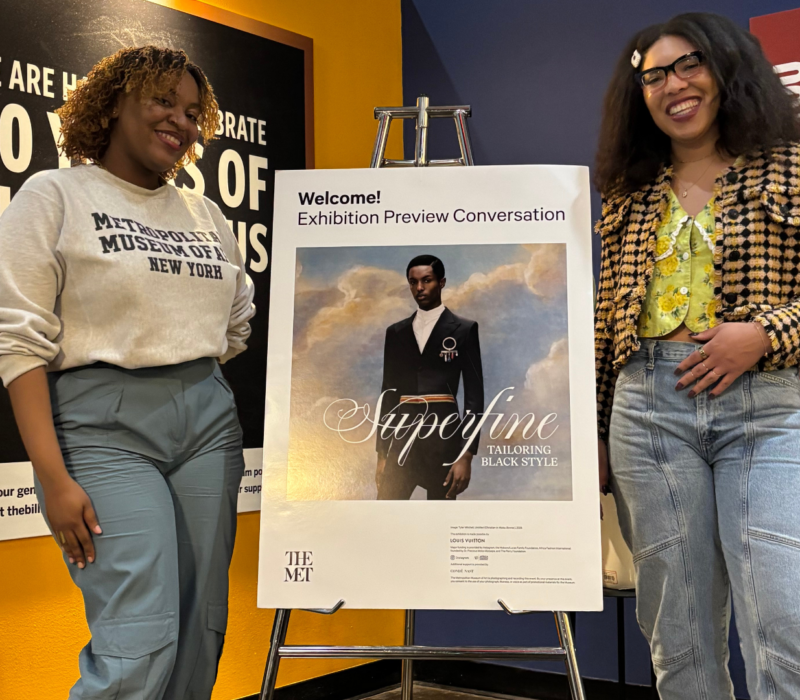 two women stand on either side of a poster advertising a new show at the Met