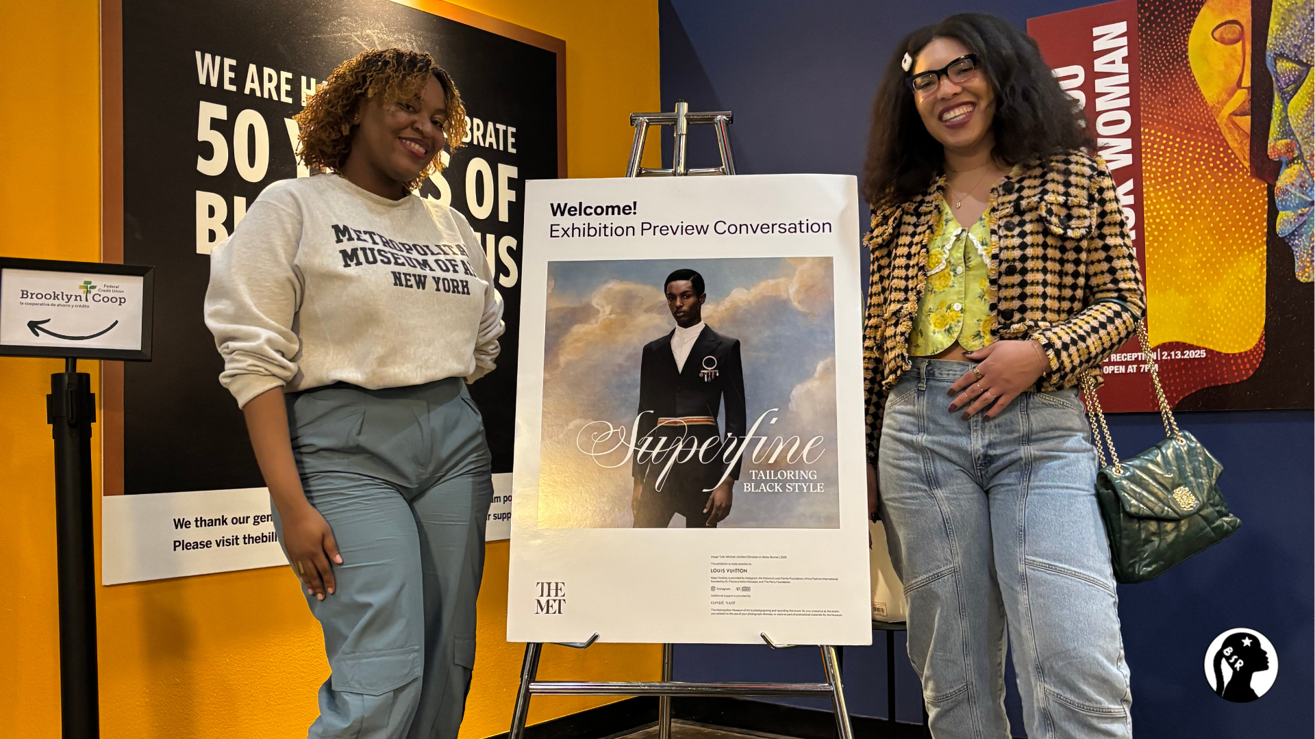 two women stand on either side of a poster advertising a new show at the Met