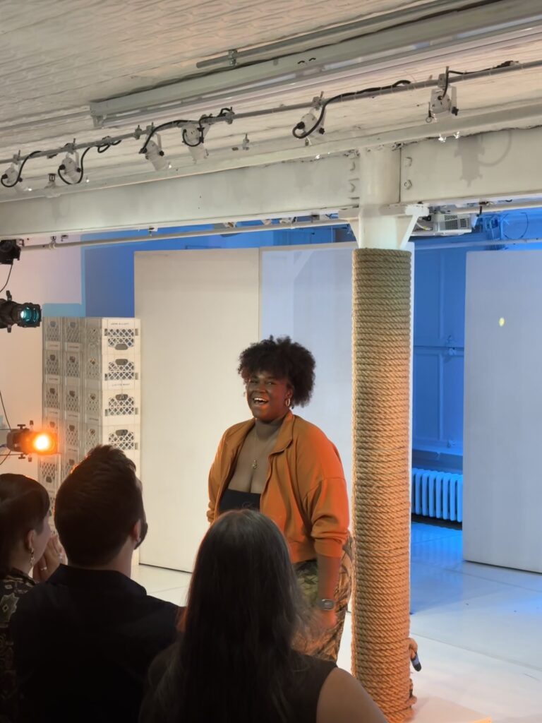 a curly-haired woman in an orange sweater smiles in front of an audience.