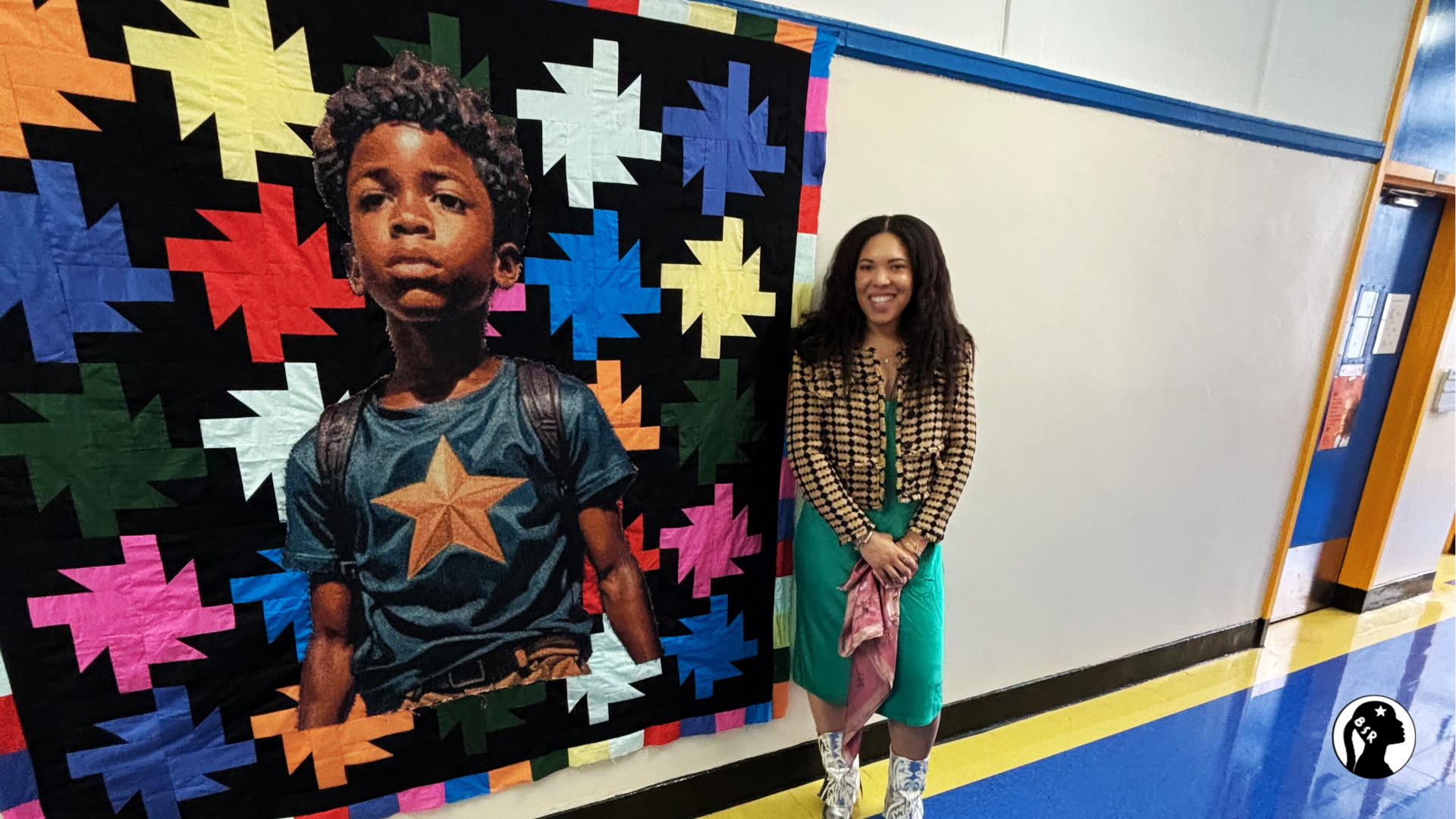 Woman stands next to a colorful quilt that features a regal Black boy.