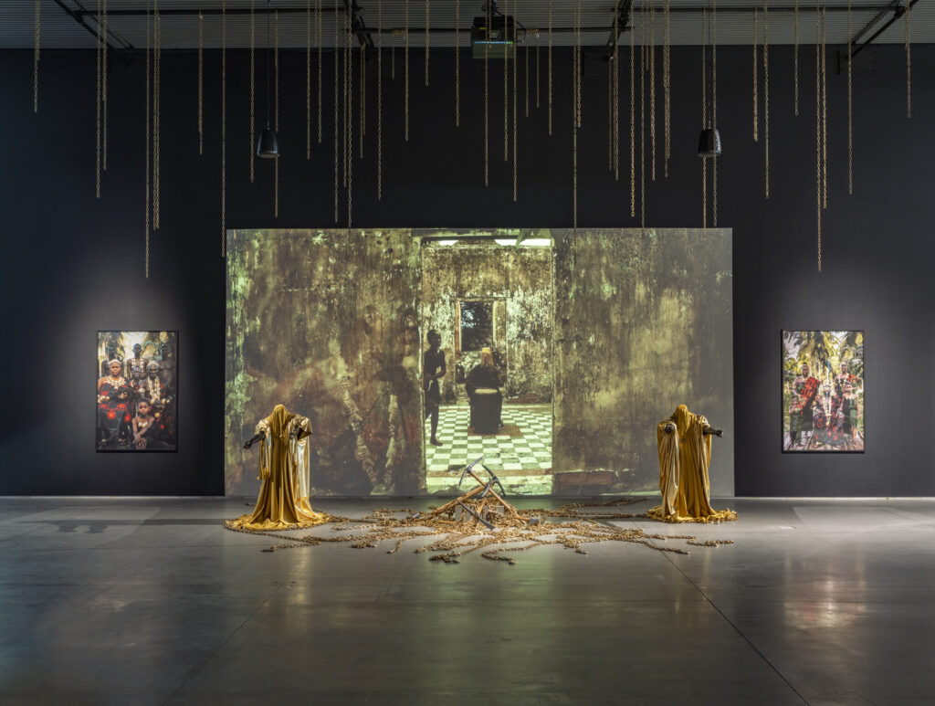 a large video screen is flanked by photographs, mannequins draped in gold, and chains hanging from the ceiling and piled on the ground