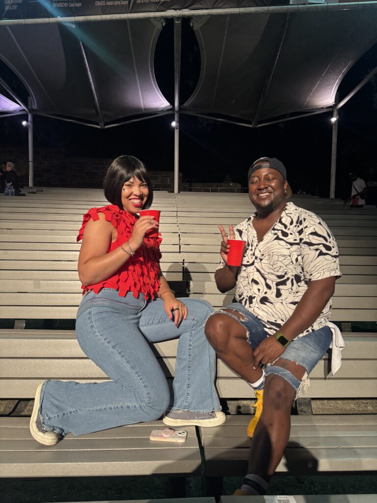 A woman in a red top and a man in a black and white button down smile in the stands