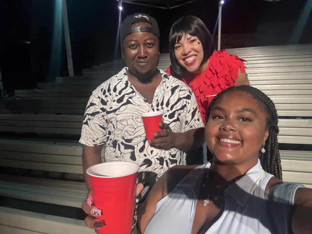A woman in a red top, a man in a black and white shirt, and a woman in a blue vest take a selfie from the stands