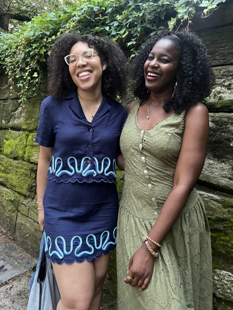 two women smile in front of a mossy wall of rocks