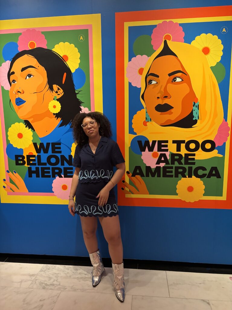a woman stands in front of two posters; one reads "we belong here" the other reads "we, too, are america"