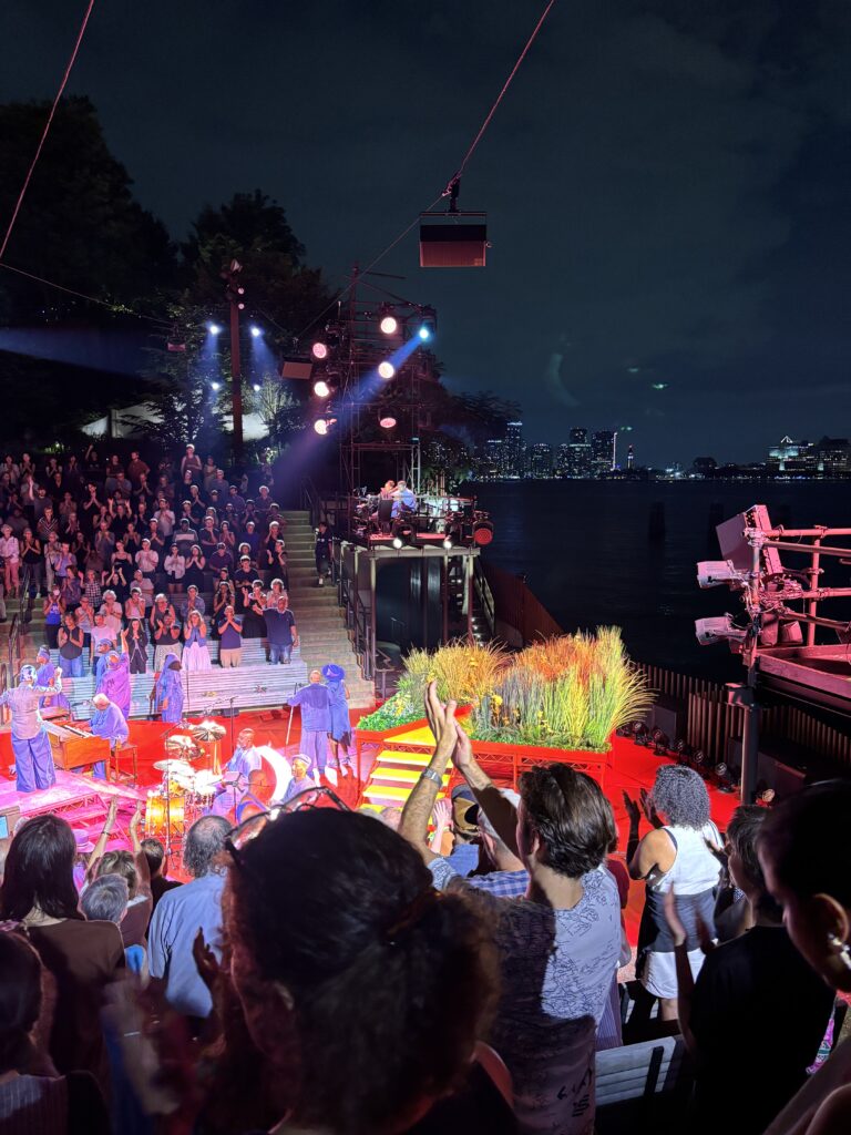 A seated crowd of people look on from their seats in an amphitheater after sunset