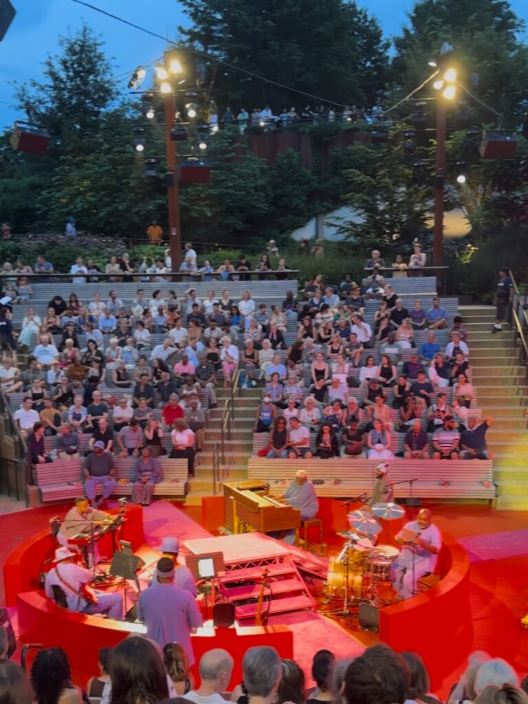 A seated crowd of people look on from their seats in an amphitheater at dusk