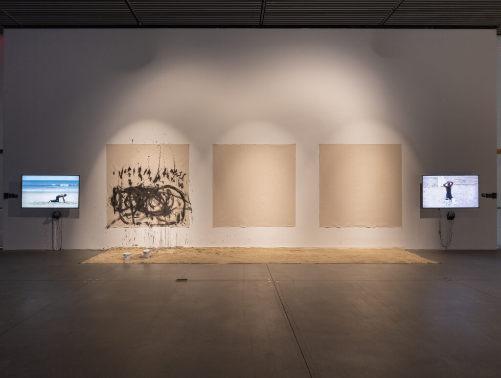 three square canvases hang on the wall between two video screens, one canvas has black paint and the other two are blank