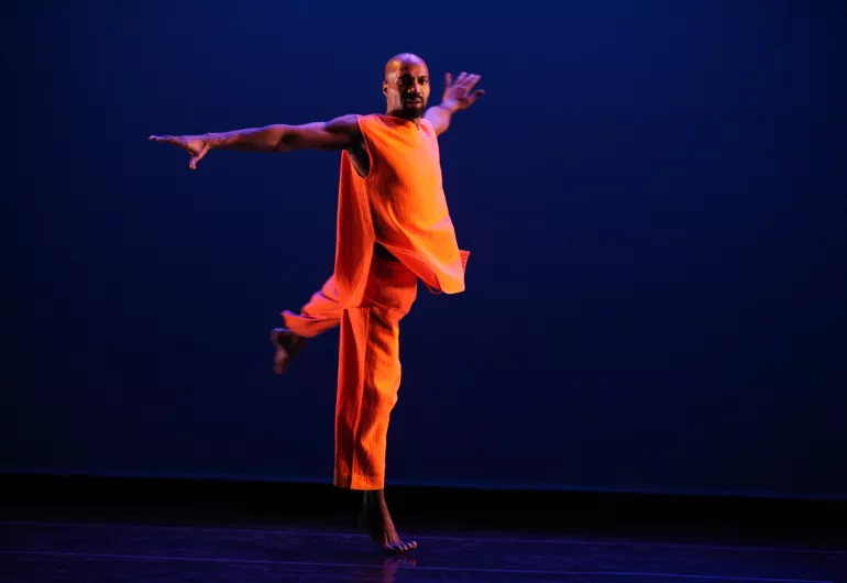 A man in a draped orange outfit stands on one foot with his arms outstretched on a stage