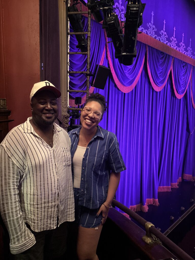 A man and woman stand in a box seats overlooking a stage with a purple curtain