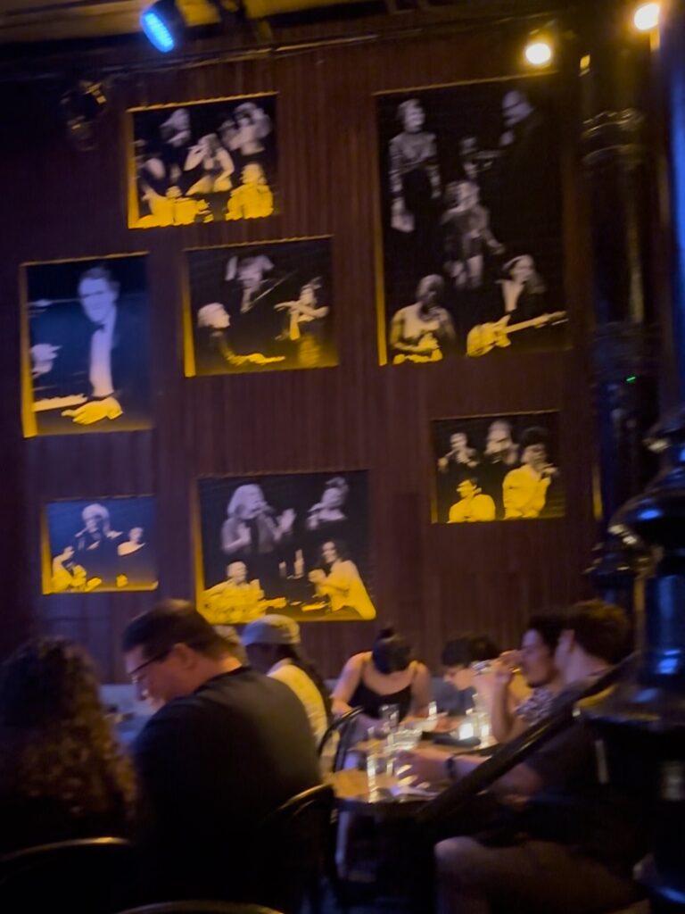 Black and white photos hang on a wooden wall. Near the base of the photos attendees huddle around tables and drinks.