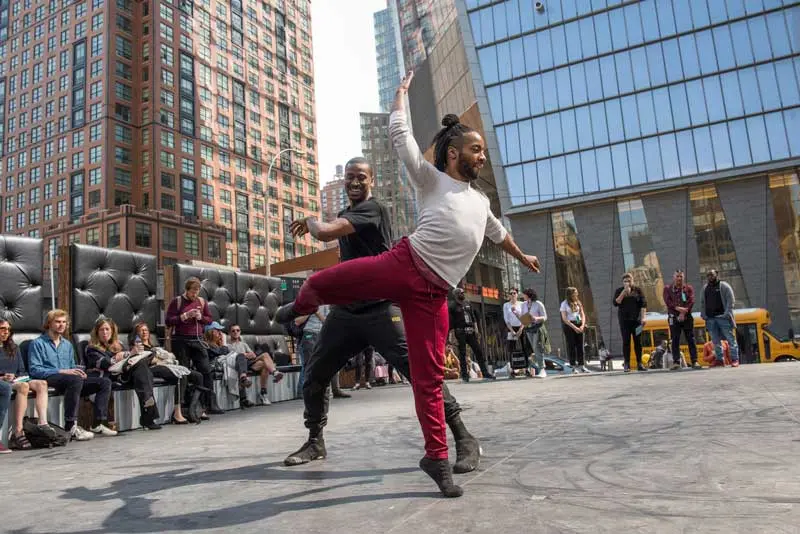 A man in a white top and burgundy pants dances on the sidewalk in front of an audience seated in a semi-circle