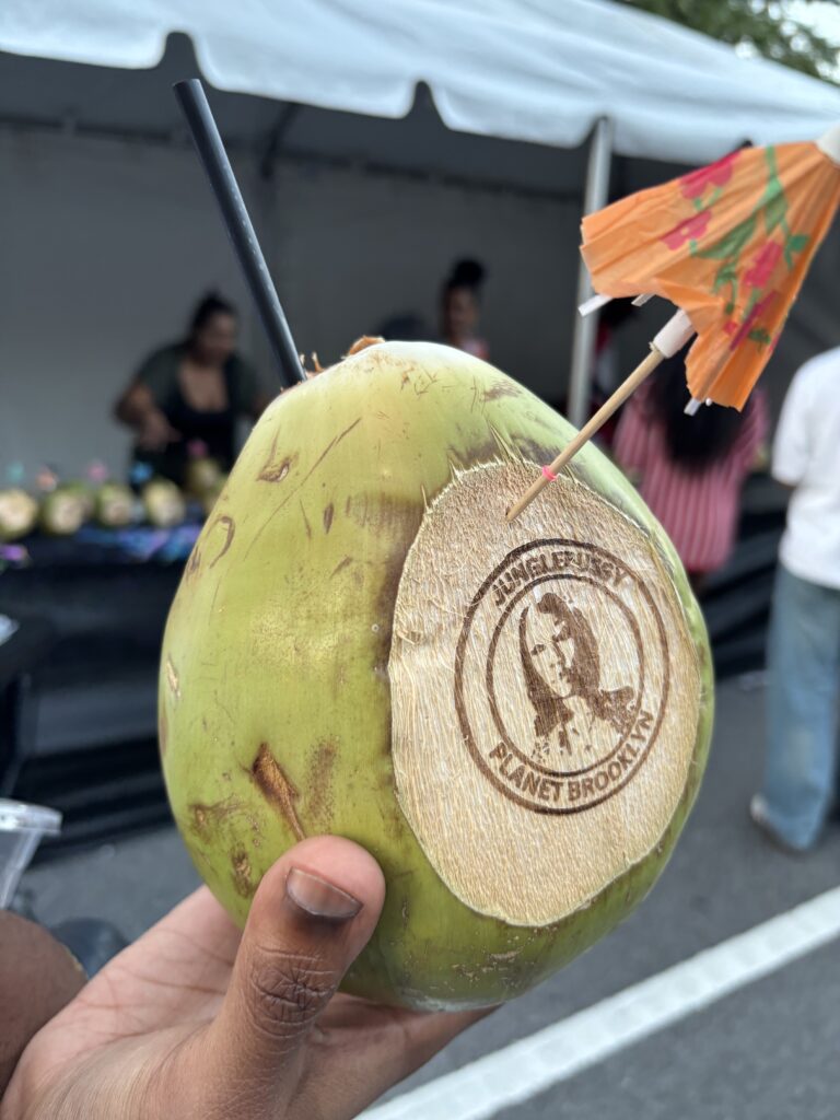 A green coconut has been shaved and branded with an iron, a straw and a orange paper umbrella stick out of it