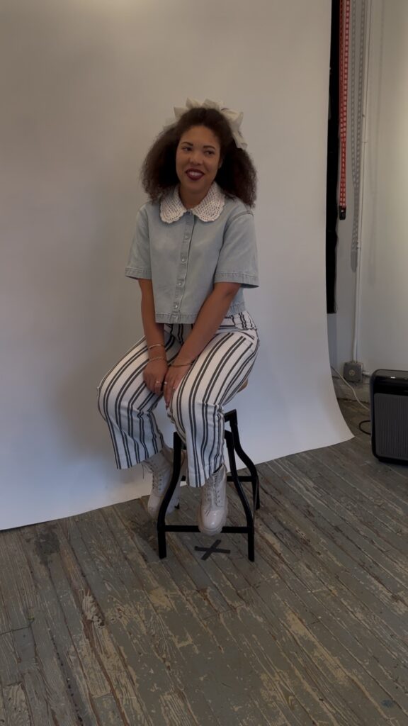 A woman in a denim collared shirt and striped pants sits smiling on a stool